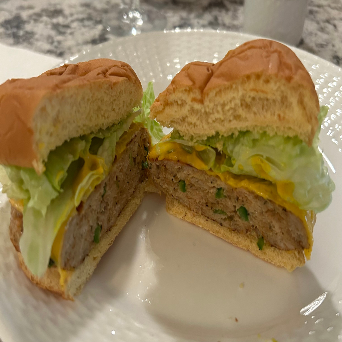 Chicken Burger cut in half on white plate. Finished product displays chicken burger on potato bun with lettuce, mustard, and melted cheddar cheese.