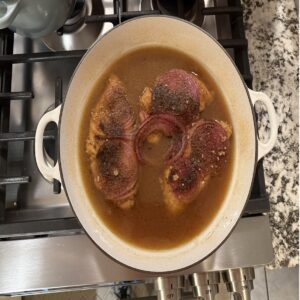 Smothered Chicken in white enamel cast iron skillet. Chicken is in beef bouillon with red onion slices on top. This is ready to serve.