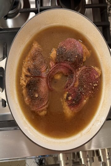 Smothered Chicken in white enamel cast iron skillet. Chicken is in beef bouillon with red onion slices on top. This is ready to serve.